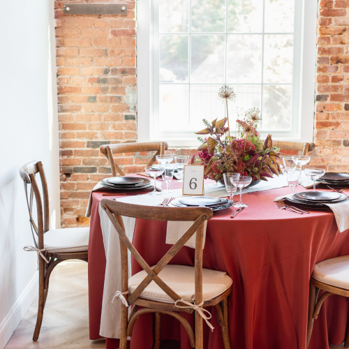 Dining table set with a copper bengaline tablecloth, floral centerpiece, and wooden chairs. Table set with black heirloom charger and dinnerware rentals. New England style gathering.